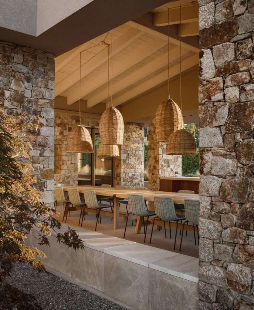 Semi-open outdoor dining area with a long wooden table, woven chairs, stone pillars, and pendant lights.