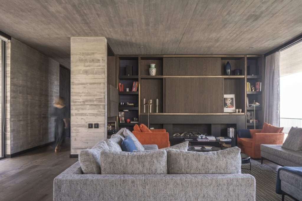 Modern interior living room in Casa Zapallar featuring board-formed concrete walls, wooden shelving unit, fireplace, and plush gray sofas.