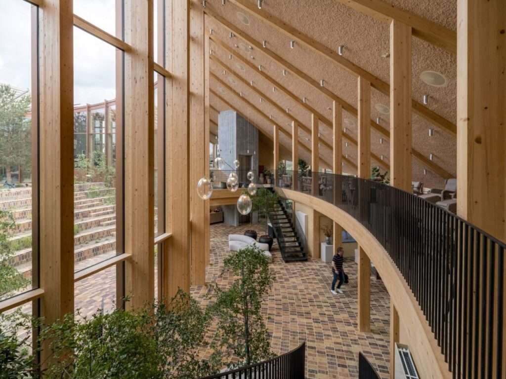 Double-height interior office atrium showing exposed mass timber columns, slanted acoustic fiber ceiling, brick flooring, and mezzanine workspace.