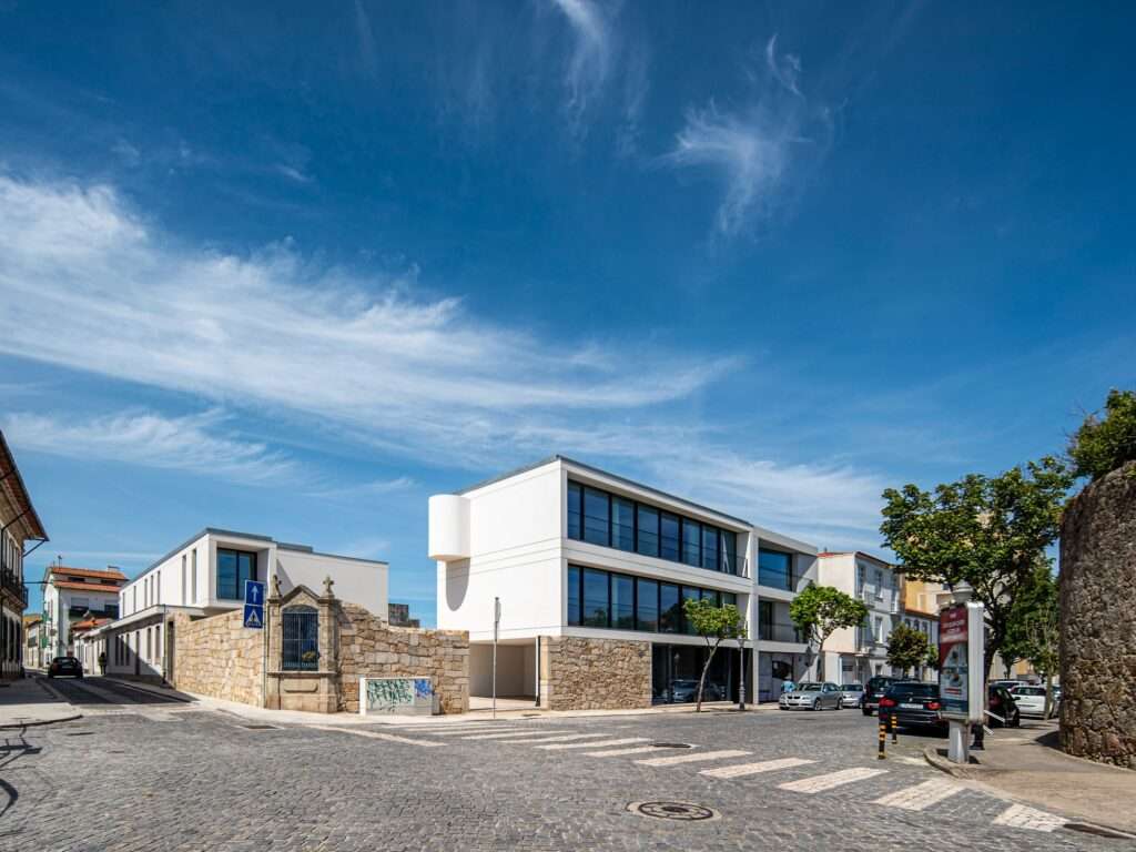Wide street corner view showing two modern white building volumes integrating existing stone walls and a historic chapel shrine in Vila do Conde.