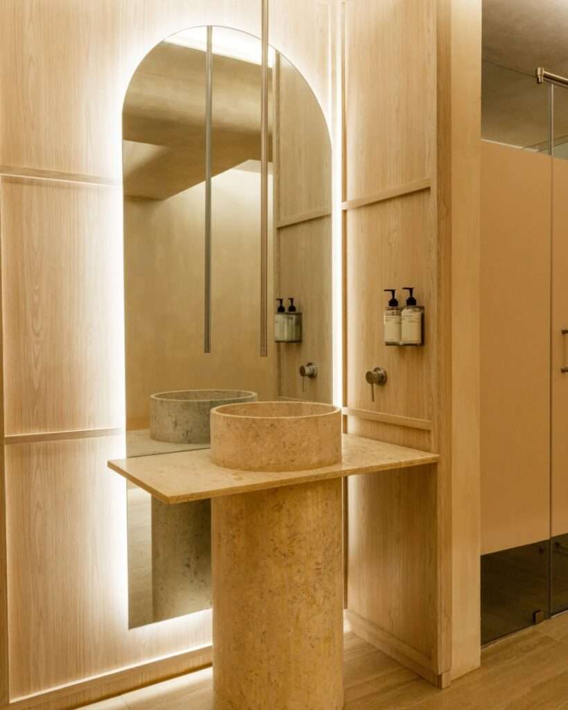 Natural travertine pedestal sink paired with an backlit arched mirror against light wood paneling.