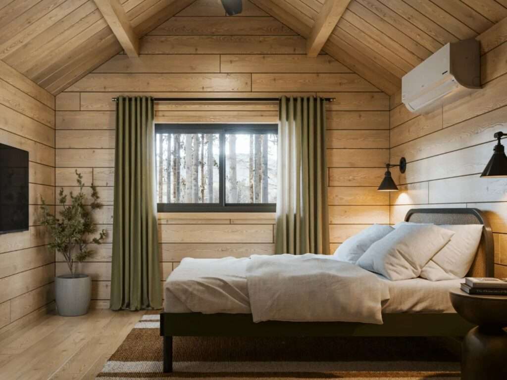 Minimalist cabin bedroom with a double bed, vaulted pine ceiling, wall-mounted air conditioner, large window with olive curtains, and black sconce lighting.