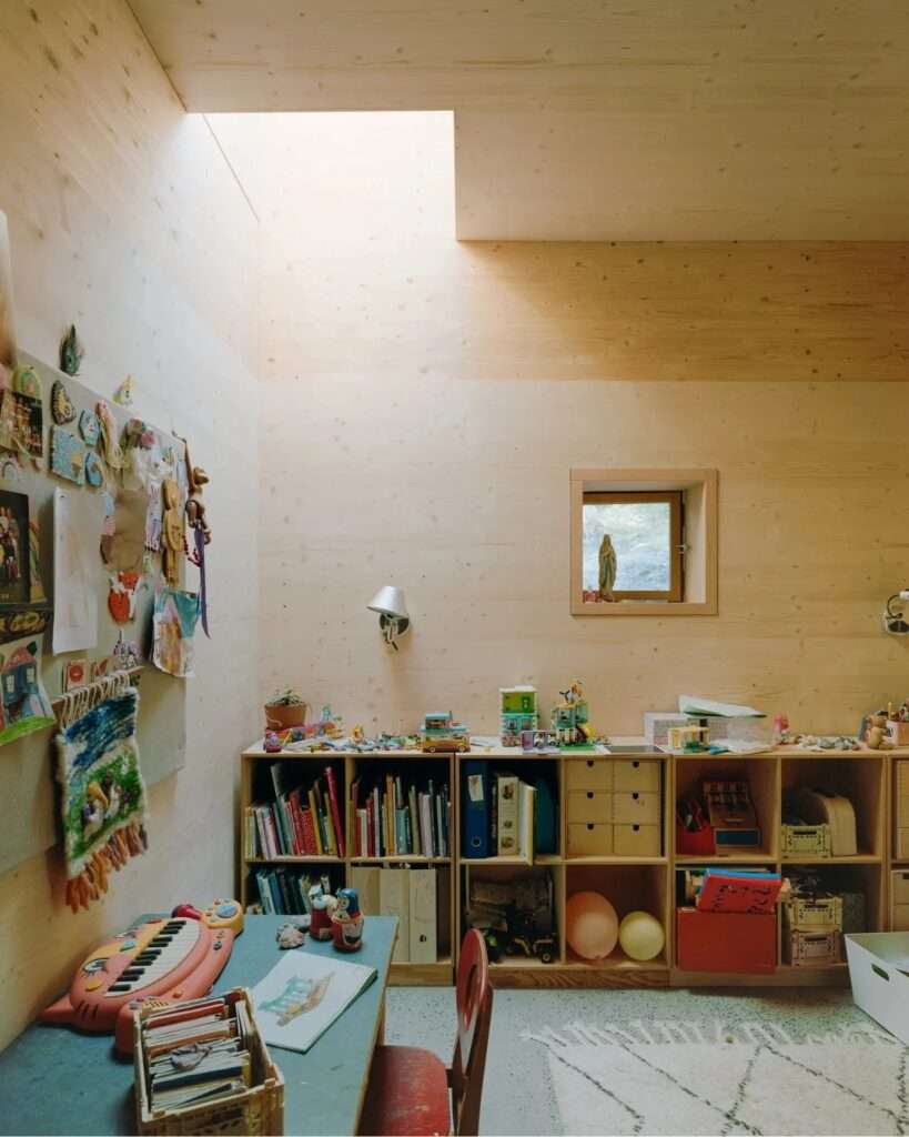 A cozy child's room or study within the wooden Ebeltoft house, featuring a large skylight, wooden bookshelves, colorful storage, a piano, and an unpainted wood ceiling and walls.