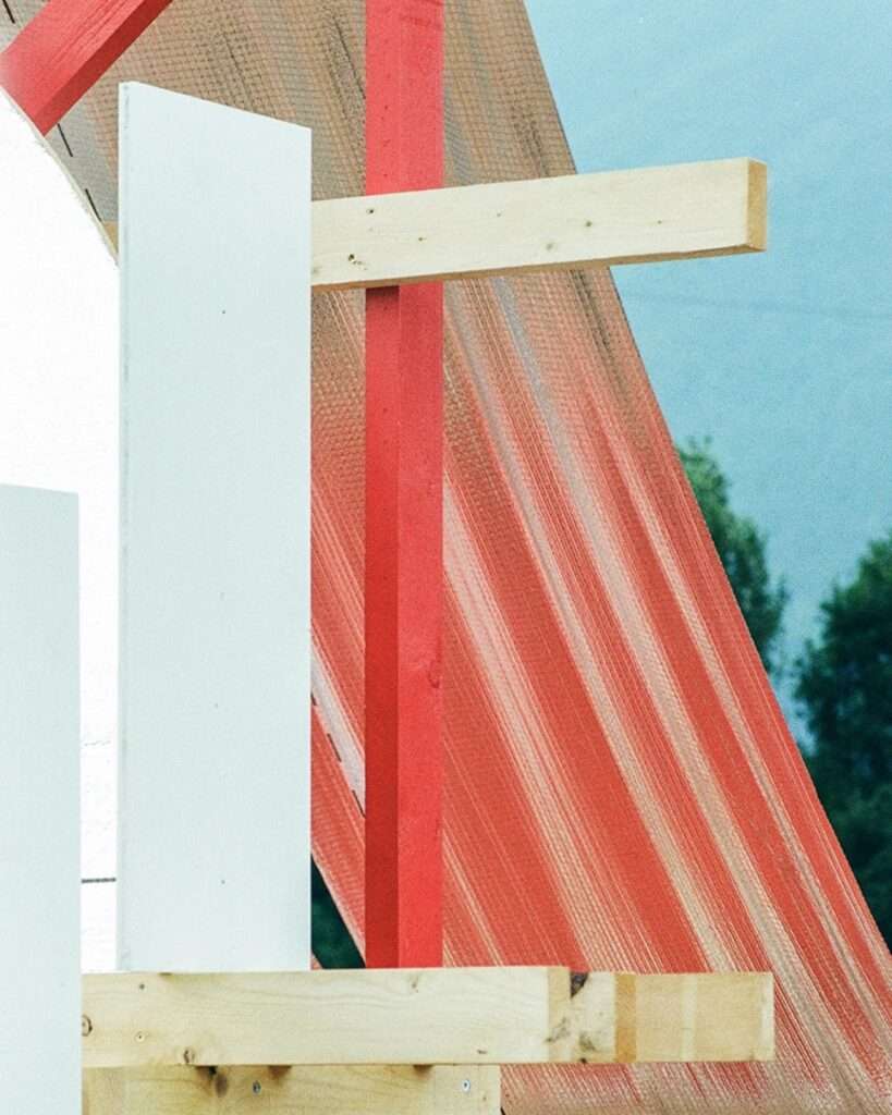 Abstract close-up of material textures on the Gabonsa Pavilion: white panels, unfinished timber, bright red painted structural elements, and reflective orange mesh against blue sky.