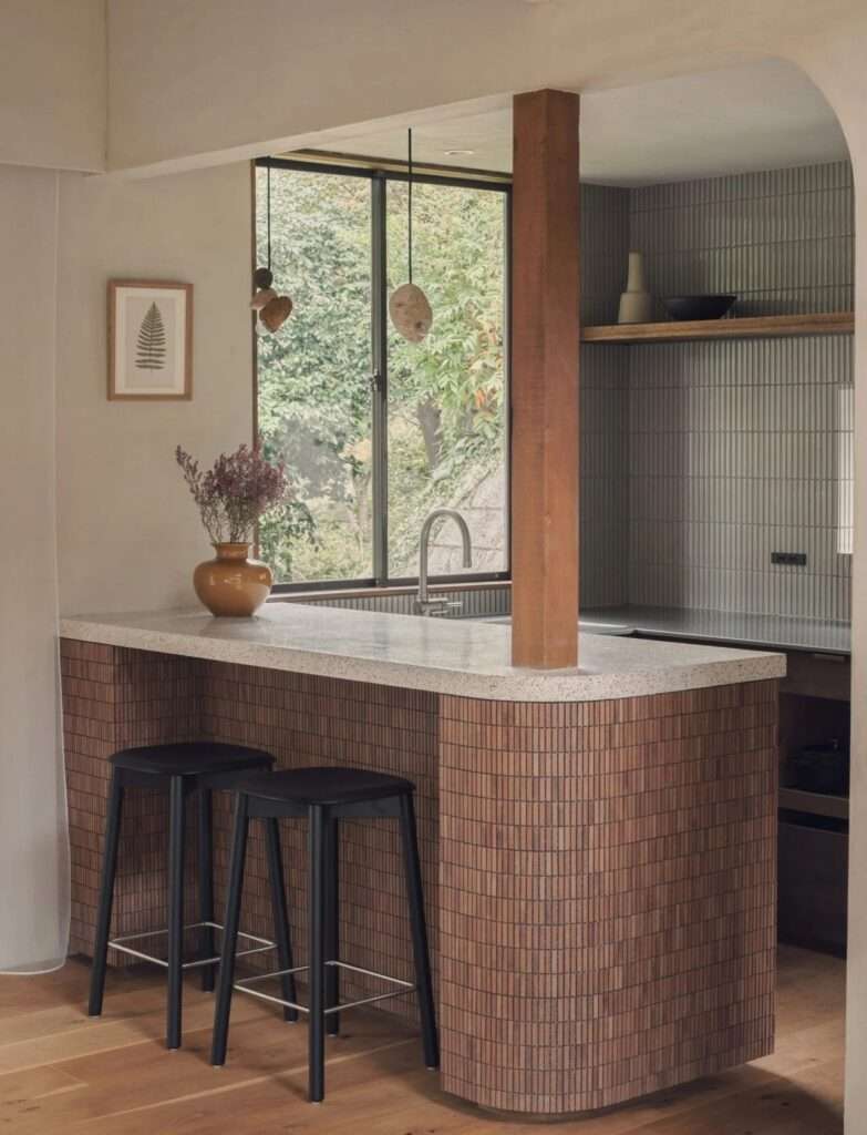 Close-up of a kitchen island clad in vertical terracotta tiles with a terrazzo countertop, black bar stools, and a timber column.