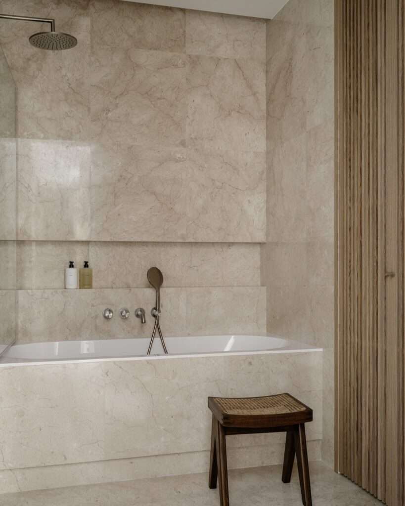 Primary bathroom with beige marble wall tiles, built-in bathtub, rainfall showerhead, and a dark wooden accent stool.