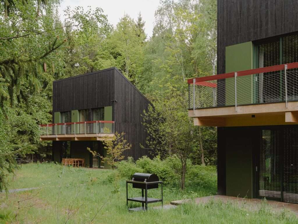 Two black timber cabins nestled in a forest clearing with a outdoor grill and green foliage.