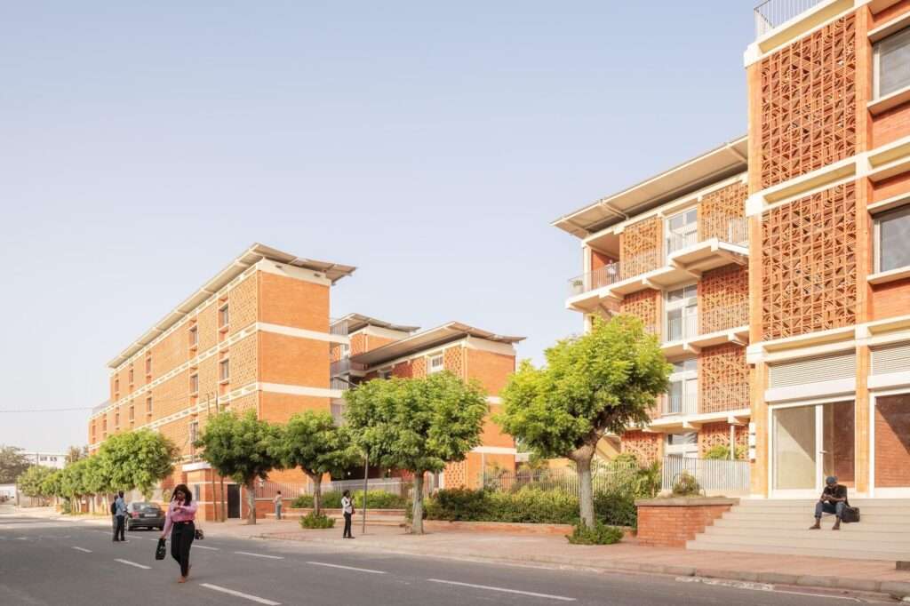 Street-level view of the four-story Cheikh Anta Diop University housing blocks clad in red brick and mashrabiya panels.