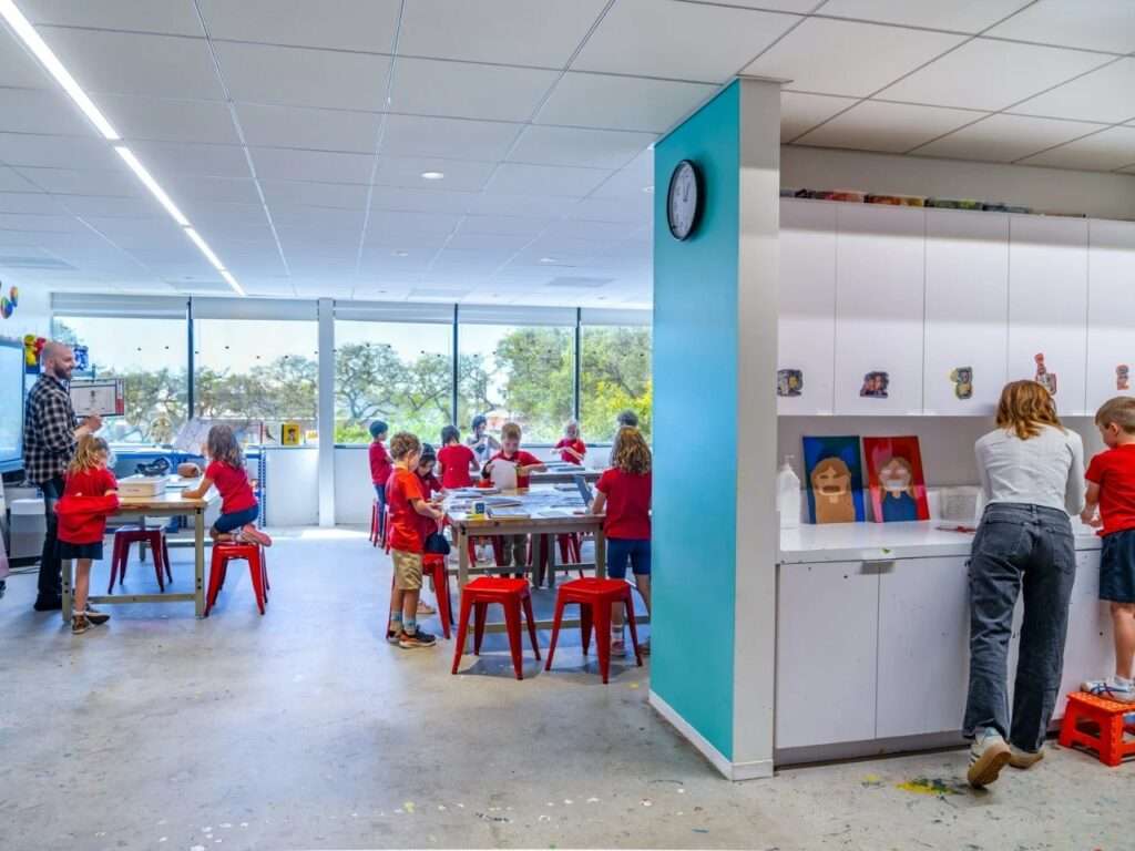 Elementary students working at art tables in a bright workshop classroom with exposed ceiling ductwork, large windows, and wall storage cabinets.