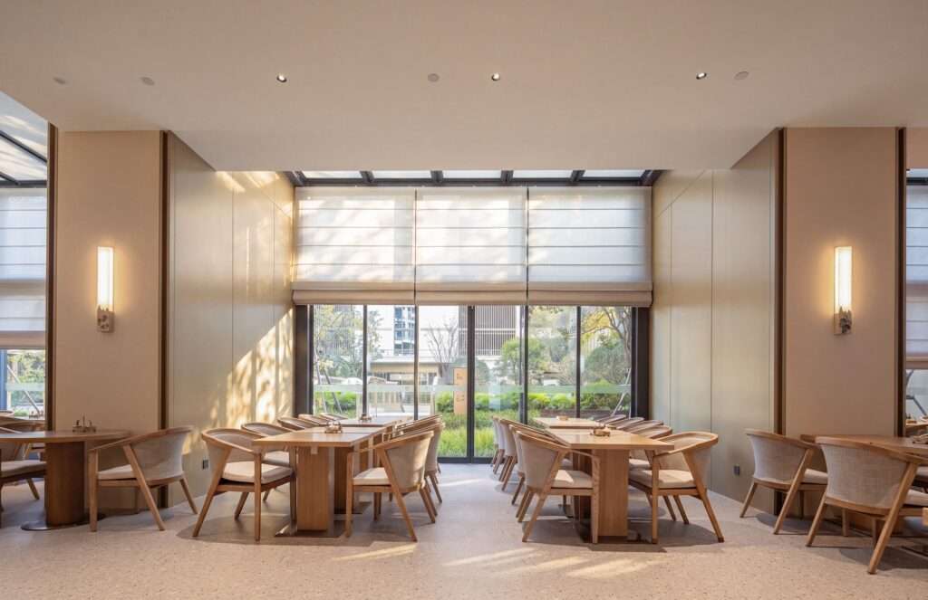 Spacious senior communal dining room with natural wood tables, comfortable cushioned chairs, and large floor-to-ceiling windows overlooking a green garden.