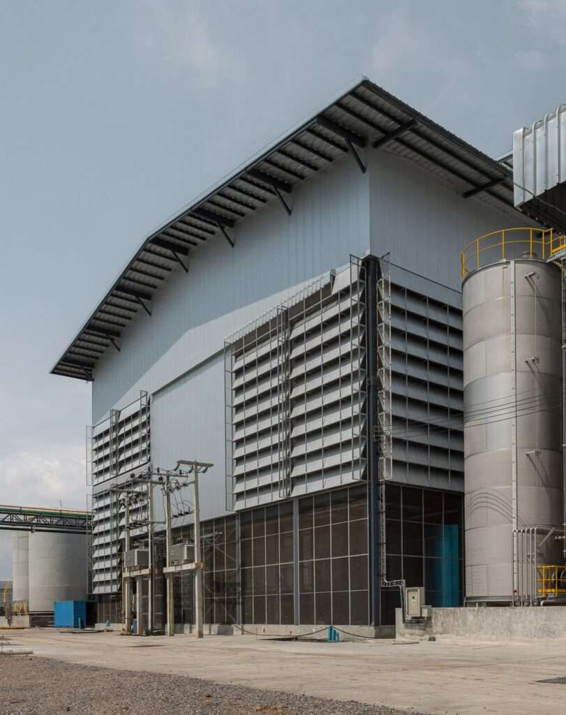 Ground-level view of the refinery building facade displaying large louvered panels, lower wire mesh enclosures, and adjacent electrical infrastructure.