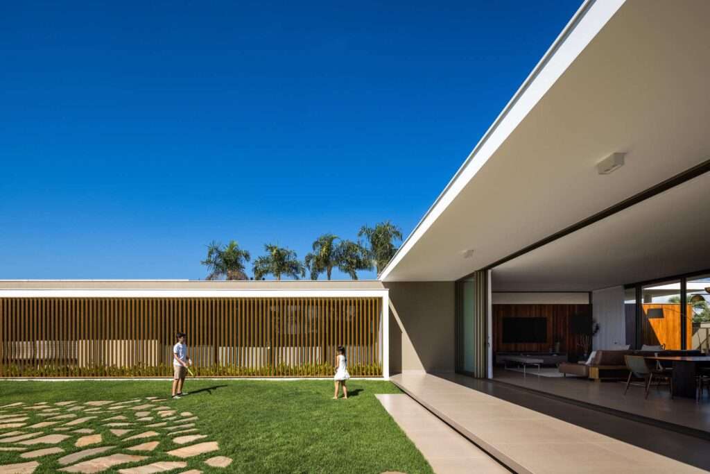 Exterior view of Refugio House showing the L-shaped cantilevered concrete roof overhang shading the open-plan social area and lawn with wooden brise-soleil louvered facade.