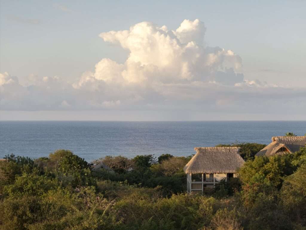 Distance landscape shot showing Casa Eva's thatched roof emerging from the lush green coastal jungle with the blue ocean and cloudy sky in the background.