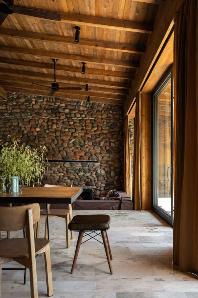 Dining room interior with wooden table, stone fireplace accent wall, floor-to-ceiling glass doors, and natural light.