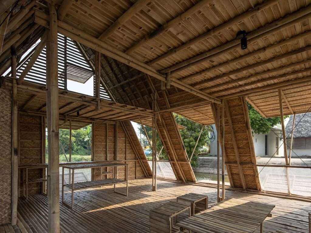 Interior shot of the Floating Bamboo House from a lower angle, showing a cluster of small bamboo stools and benches on the textured woven floor, with light and shadow cast by the extensive open bamboo roof.