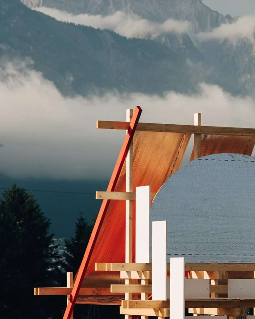 Close-up detail view of the Gabonsa Pavilion, showing the highly reflective orange material, white panels, and wooden beams against a background of mountains and dark trees under a hazy sky.