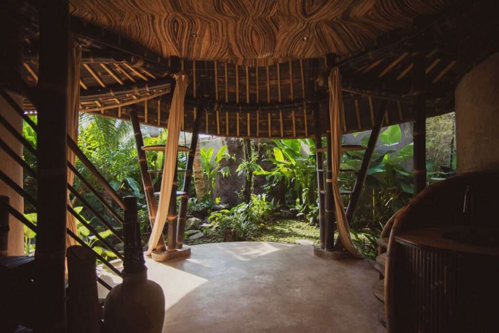 Interior view looking out towards a lush tropical garden through an open circular bamboo structure with hanging drapes, highlighting the seamless indoor-outdoor transition and a large earthenware pot.