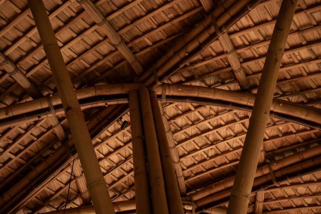 Low-angle interior detail of intersecting bamboo columns, structural joinery, and woven thatch roof lining.