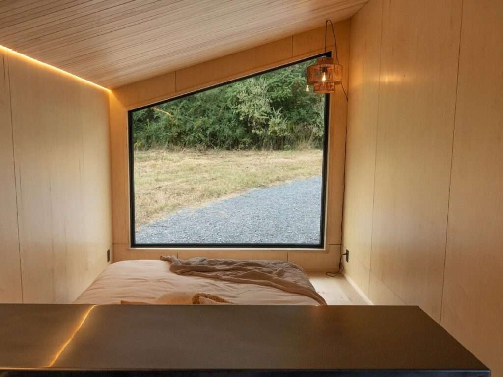 View from inside the Rogue Mini mobile home looking out through a large, panoramic window toward a field and treeline. The walls and ceiling are lined with light-colored wood panels. LED strip lighting glows along the ceiling fold.