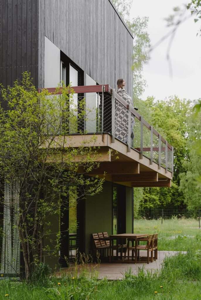 Side view of a black cabin balcony with a person standing on the deck wrapped in a blanket, looking out at the forest.
