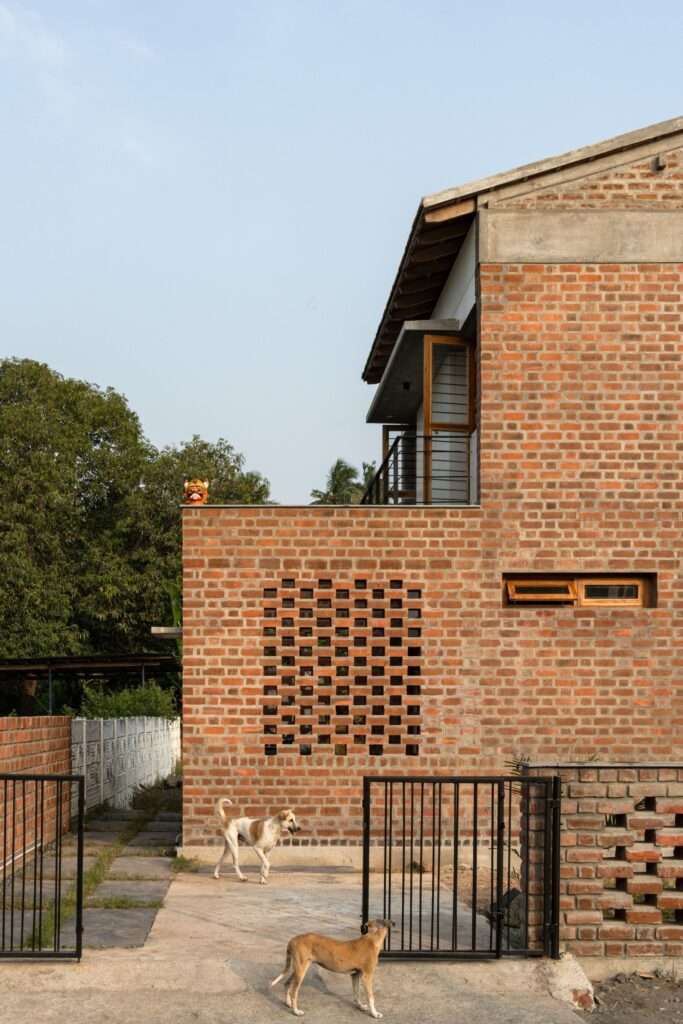 Exterior front facade of Thuraiyur Farmhouse, featuring exposed brick walls with a square jali opening, an iron entry gate, and local dogs standing on the paved entryway.