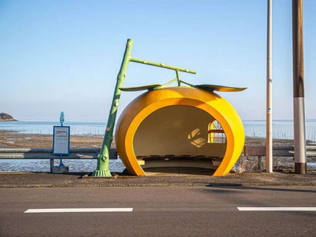 Tangerine orange fruit-shaped bus stop with a green stem support structure along a seaside road.