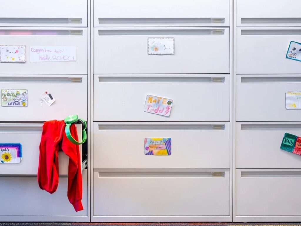 White lateral file cabinet drawers adapted into student storage, decorated with personalized student name tags, drawings, and a red hoodie.