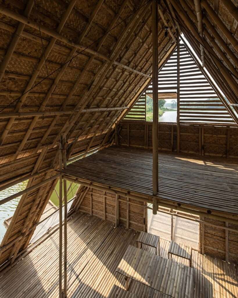 Interior of the Floating Bamboo House, looking from a mid-level platform or loft, straight towards a window at the peak of the triangular gable, revealing a high-set triangular opening with louvers and a view of trees.