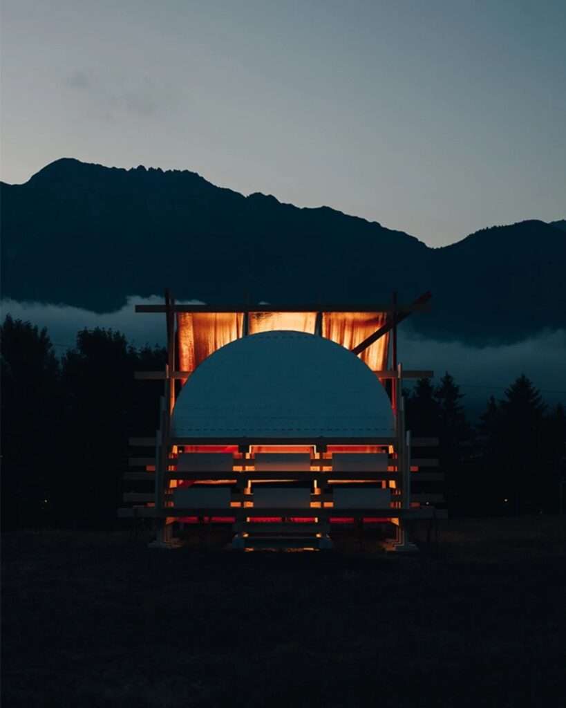Exterior view of the Gabonsa Pavilion at dusk. The interior is warmly illuminated, casting a glow on the reflective orange mesh and the white semi-circle. Mountains and trees are in deep shadow under a twilight sky.