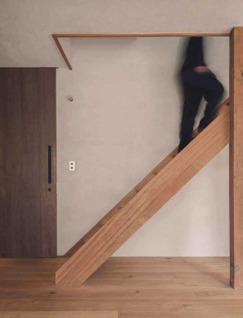 Minimalist wooden staircase against a neutral plaster wall with a silhouette of a person ascending the steps.