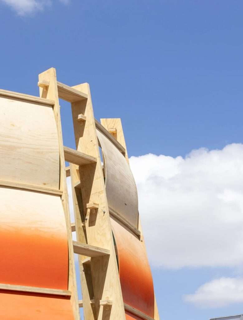 Low-angle shot of curved orange plywood panels and wooden ladder-like frame against a clear blue sky.