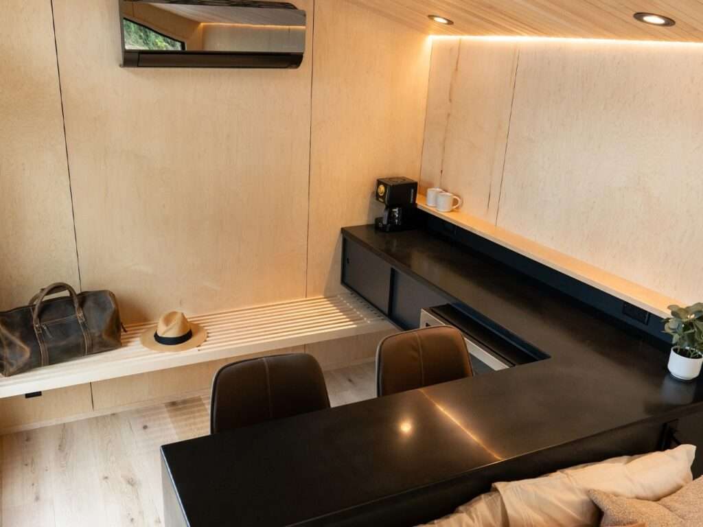 A view of the internal organization of the Rogue Mini, showing a black countertop, integrated workspace with seating, a small refrigerator, a ducted air conditioning unit on the wood-paneled wall, and a bench with travel items (hat and bag).