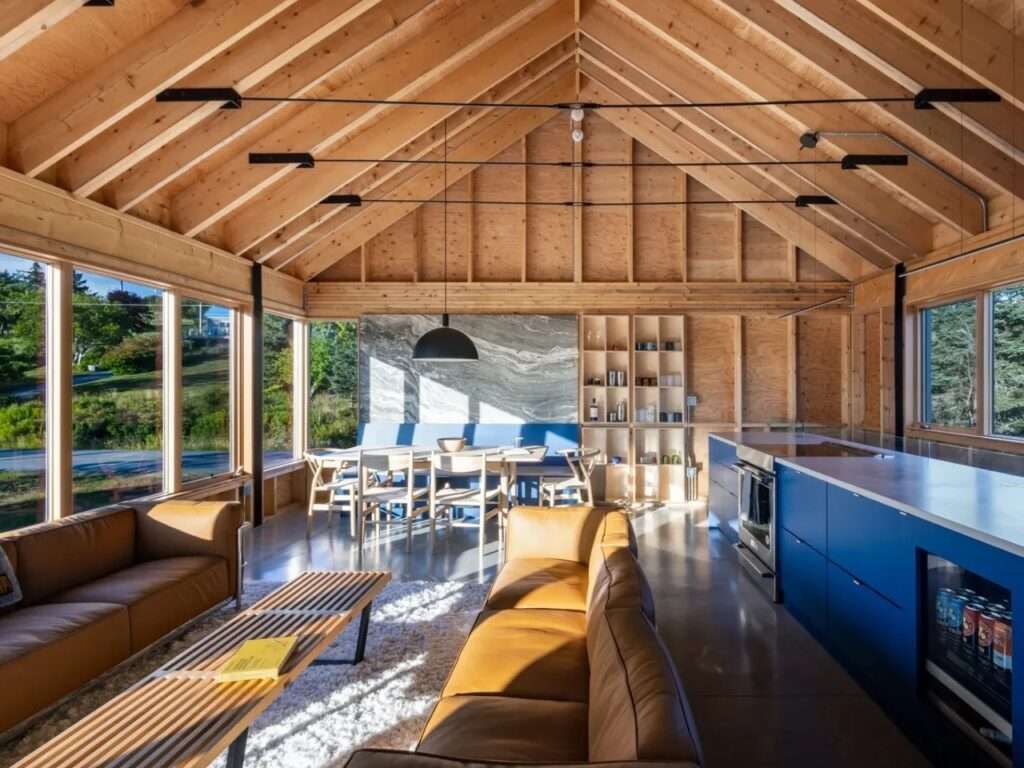 Open-plan upper floor of Skybox featuring vaulted timber roof structure, blue kitchen island, leather seating, and surrounding ribbon windows.