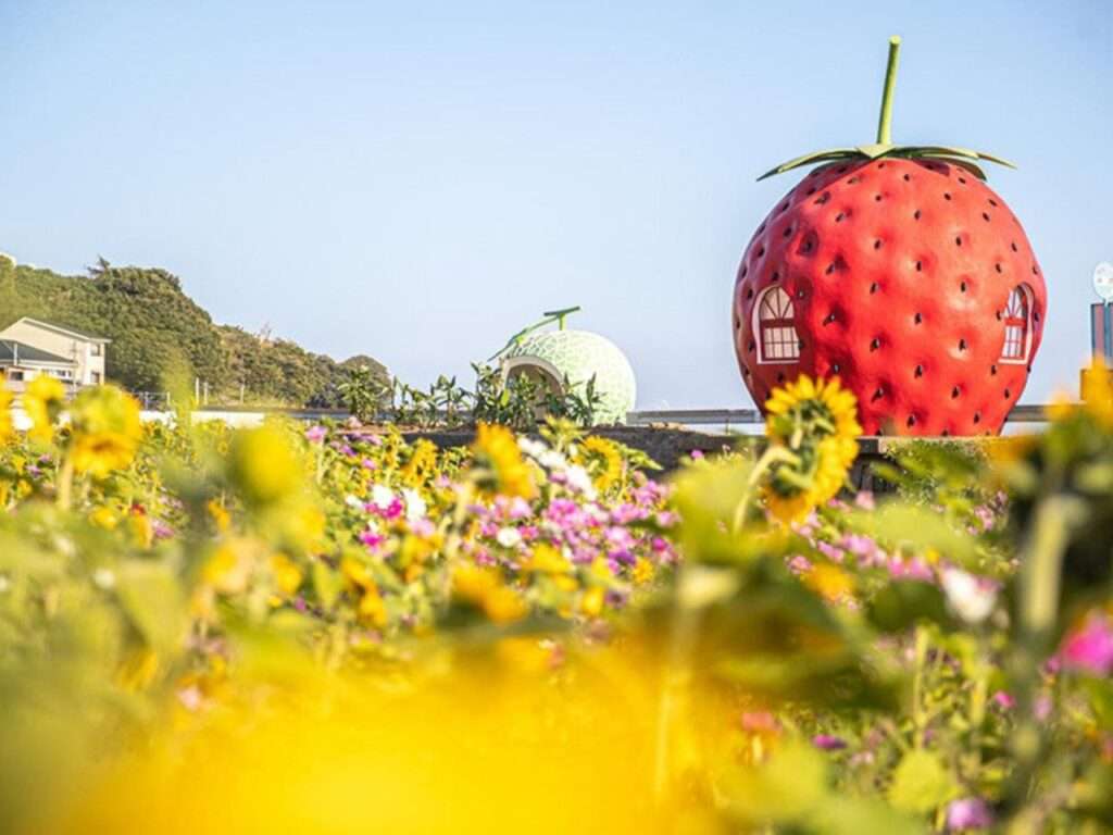Giant strawberry-shaped bus shelter with detailed seed textures standing near a flower-filled field.