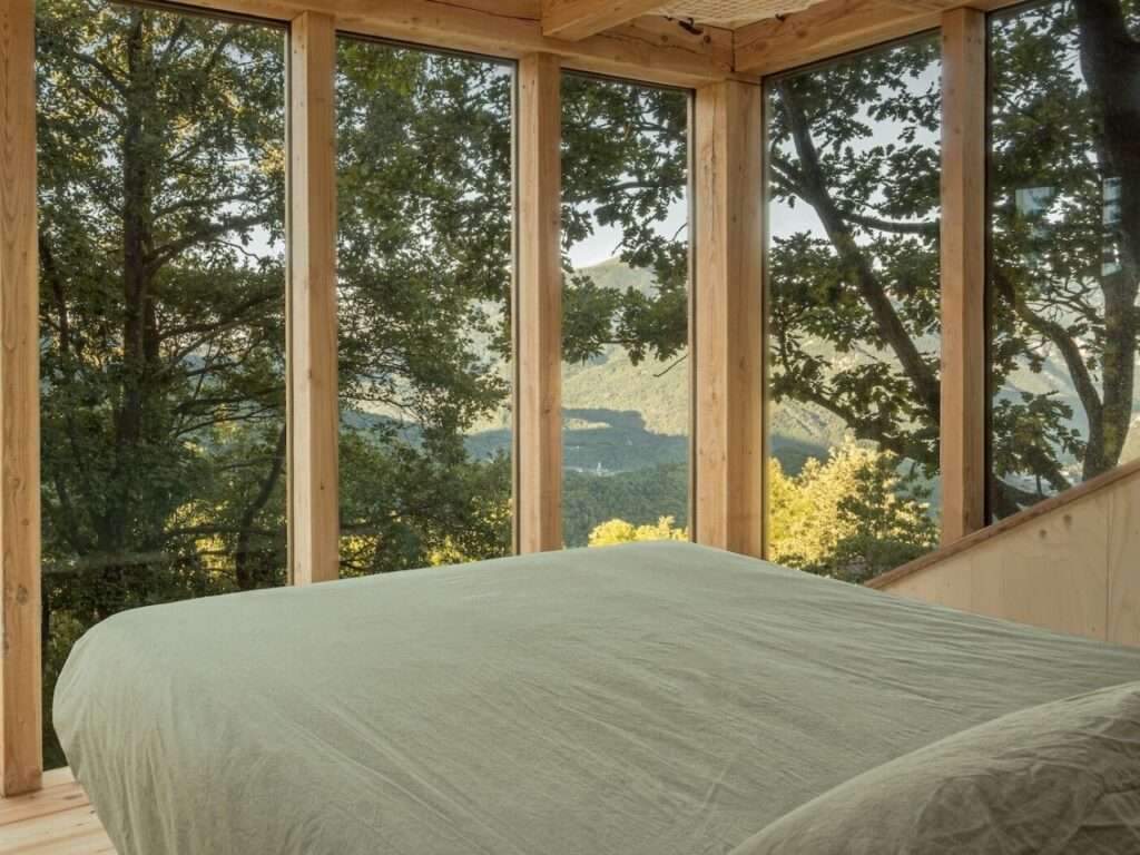Bed in TreeBOX cabin facing floor-to-ceiling glass window overlooking forest and alpine valley.