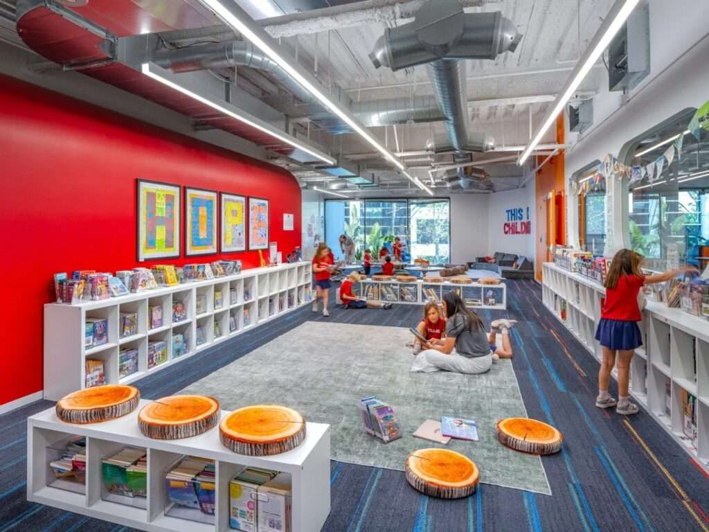 Reading library space with low bookshelf units, tree-stump seating cushions, red feature wall with letter artwork, and students reading on a central rug.