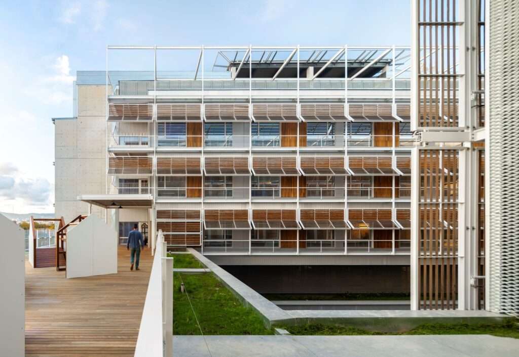 Courtyard view of Hospital del Mar expansion showing timber solar louvers, balconies, and an outdoor courtyard walk.
