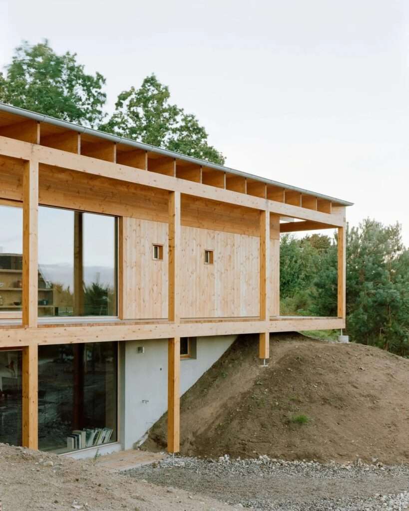 Close-up of the House in Ebeltoft showing the modular wooden structure, exposed roof beams, large glass windows, and concrete lower level anchored into a dirt and gravel slope.
