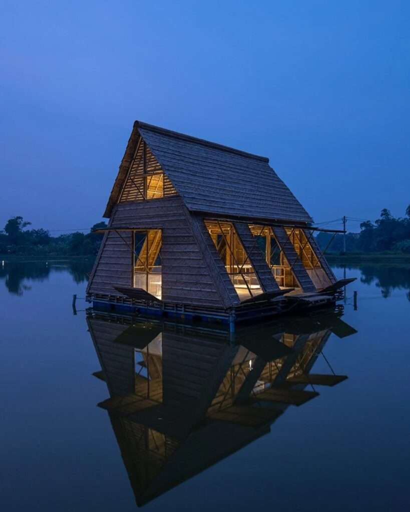 Night view of the Floating Bamboo House, with warm internal light glowing from every opening, its reflection perfectly mirrored on the dark water of the Mekong Delta, under a deep blue sky.