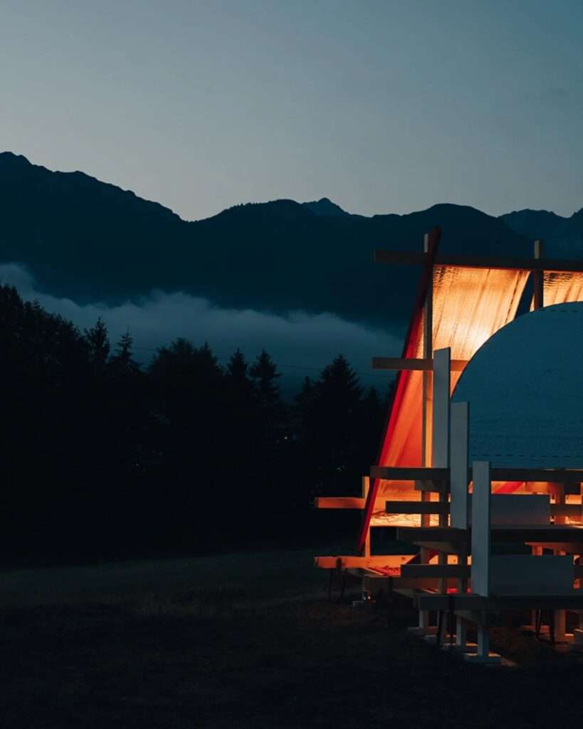 A close-up night view of the illuminated Gabonsa Pavilion. Warm light glows through the reflective orange mesh and behind the semi-circular dome, highlighting the structural wooden frame against dark mountains and trees.