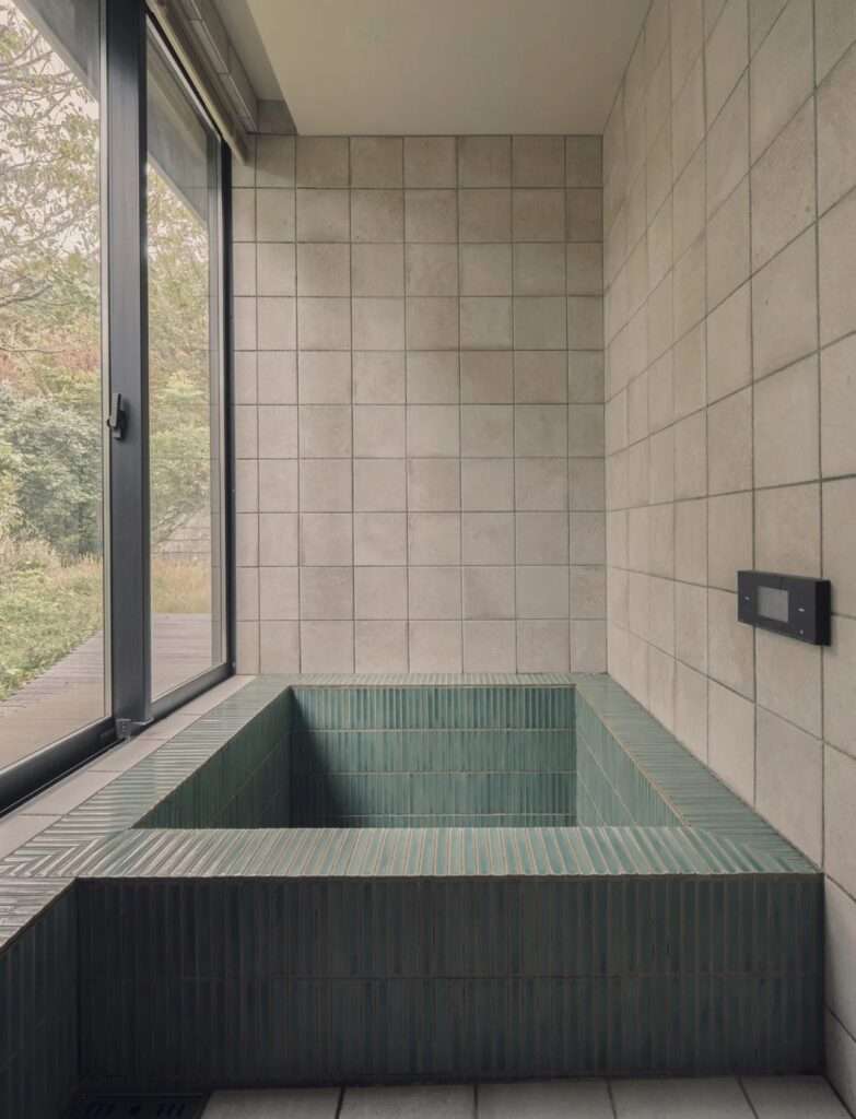 Sunken Japanese soaking tub lined with teal vertical tiles next to a floor-to-ceiling glass window overlooking nature.