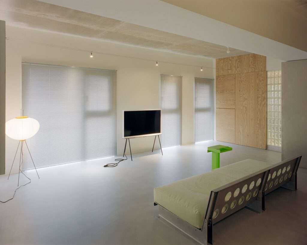 Minimalist living room with easel-mounted television, paper tripod floor lamp, window blinds, exposed concrete ceiling, and fluorescent green side table.