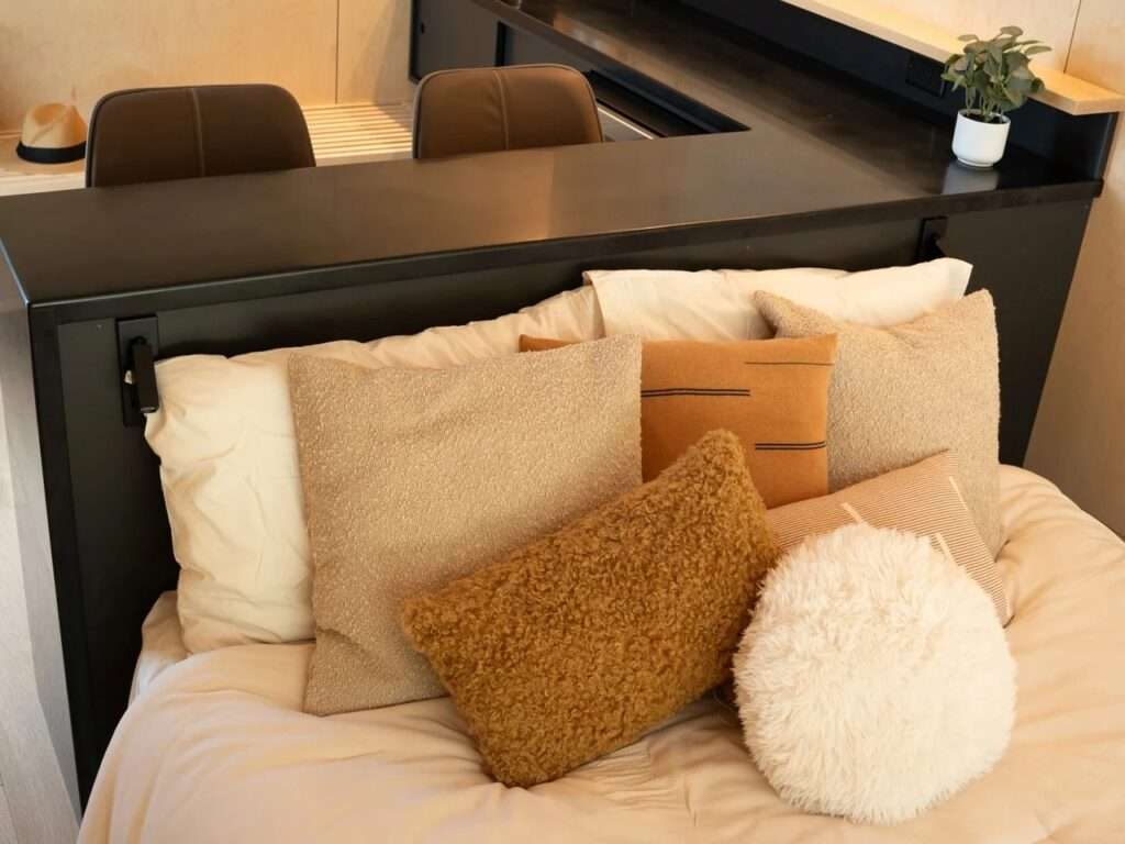 Close-up of the bed area in the Rogue Mini, showing various textures of bedding, including wool and fuzzy pillows in neutral earth tones, against a light wood panel backdrop. The smooth black countertop element frames the foreground.