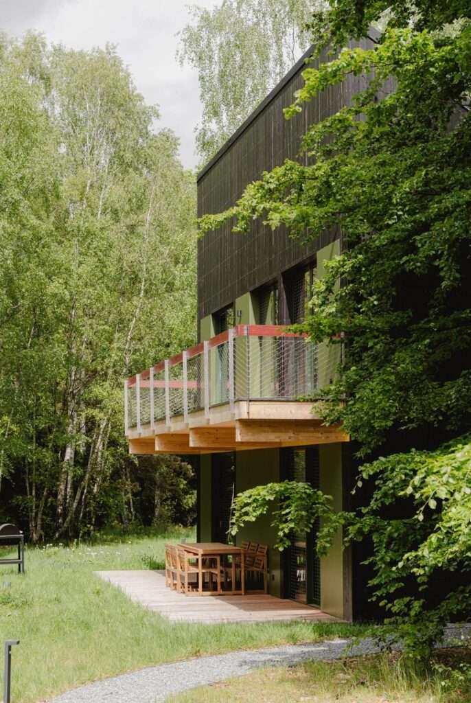 Ground floor wooden terrace with outdoor table and chairs beneath a second-story cantilevered balcony surrounded by trees.