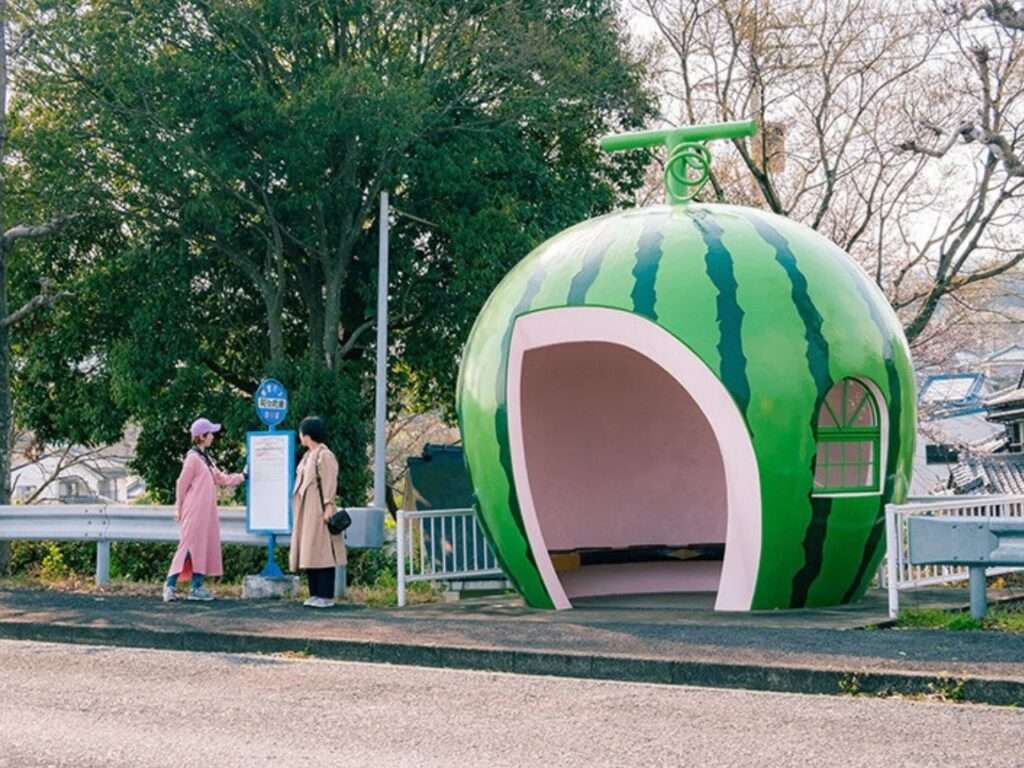 Watermelon-shaped bus stop shelter with green stripes and passengers standing near the bus sign post.