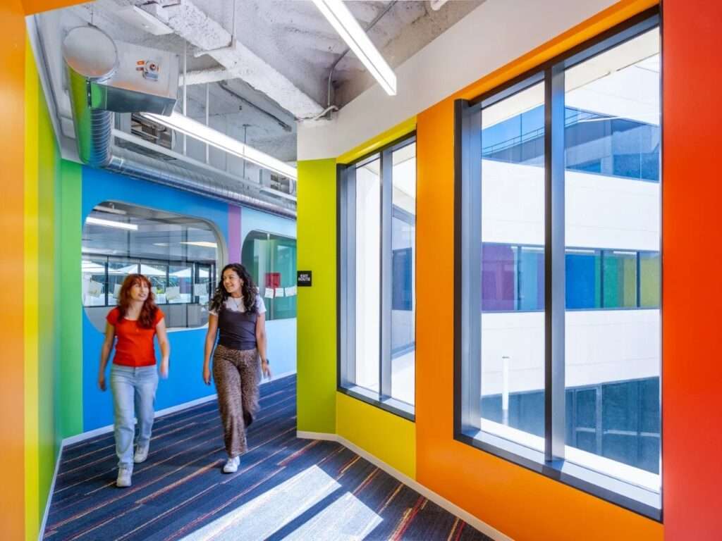 Two instructors walking down a colorful green and orange interior corridor lined with large windows overlooking a central exterior courtyard.