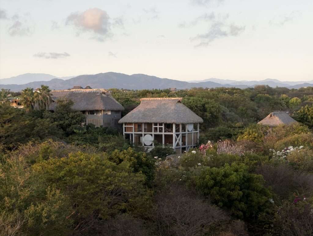 Wide aerial perspective of Casa Eva surrounded by dense forest foliage, neighboring thatched buildings, and mountain ranges under a light sky.