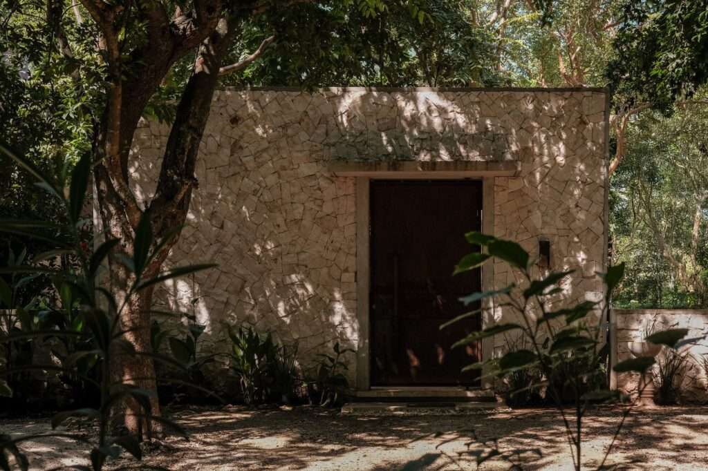 Free-standing stone masonry pavilion with dark entrance door surrounded by tropical trees and dappled sunlight.