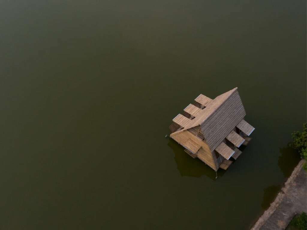 High aerial perspective looking straight down upon the entire exterior of the Floating Bamboo House, showing its triangular thatched roof, the platforms extending from all sides, and the grid of blue barrels below, floating on the vast expanse of murky water.