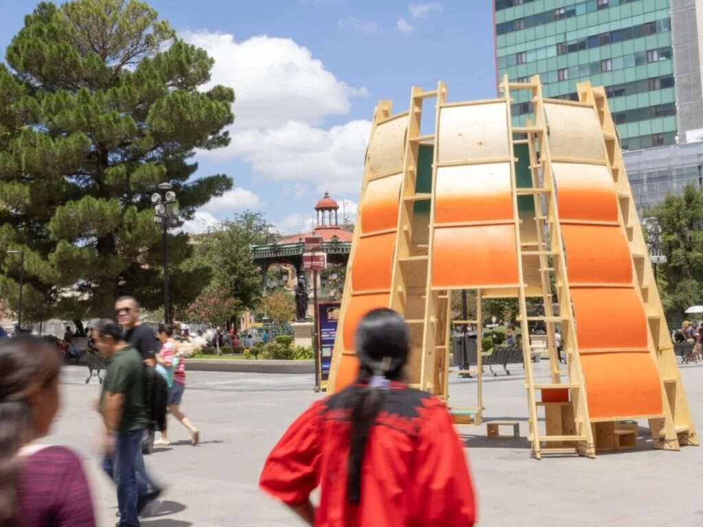 Motion blur of pedestrians walking past the bright orange Paso Nara architectural pavilion in the square.